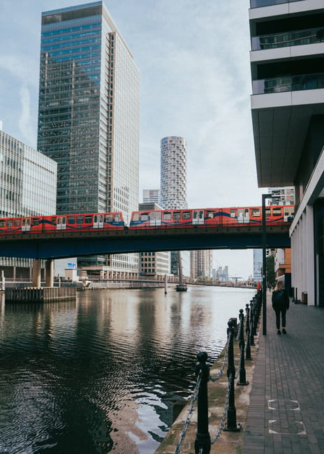 This image depicts a canal scene in Canary Wharf featuring modern high-rise office buildings with glass facades reflecting daylight. A bright red London Underground train crosses a bridge over the wat