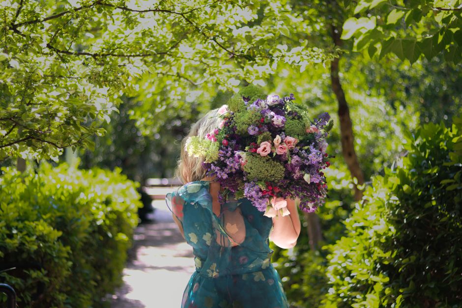 A person holding a large, vibrant bouquet of fresh flowers in front of their face, standing outdoors on a sunlit pathway surrounded by lush green foliage and trees. The bouquet features a mix of pink,