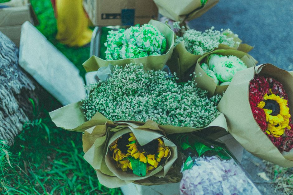 A variety of fresh flower bouquets arranged with colorful carnations, baby's breath, and sunflowers, wrapped in brown craft paper and displayed on a surface outdoors. The bouquets feature vibrant reds