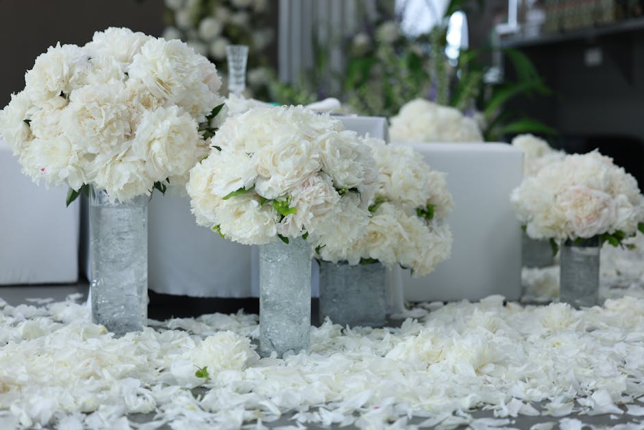 Elegant floral arrangement featuring large, fluffy white hydrangea and peony bouquets placed in tall, cylindrical glass vases with a frosted appearance. The flowers are fresh, with tightly packed peta
