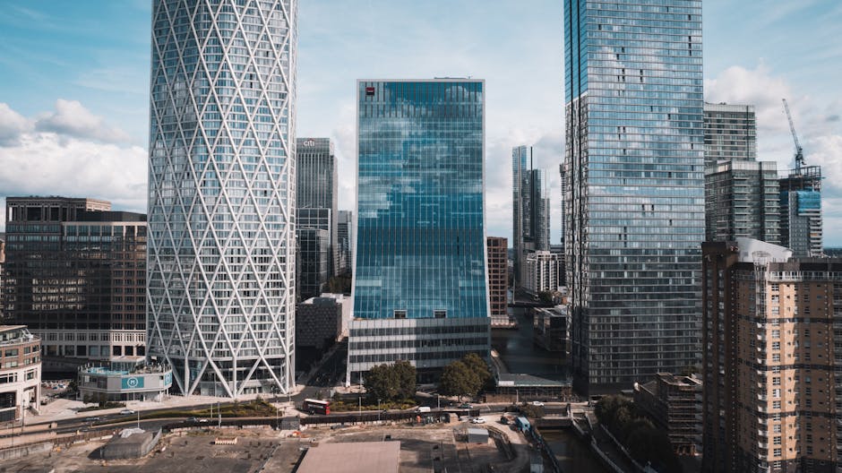 A cluster of modern skyscrapers in Canary Wharf, including the distinctive twisting design of the One Canada Square tower, with glass exteriors reflecting the partly cloudy sky. The buildings vary in 